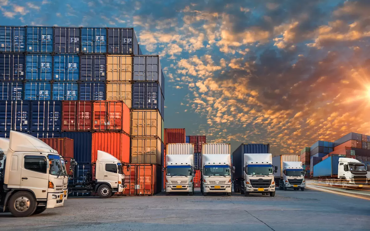 Trucks parked in front of stacked shipping containers at a logistics terminal during sunset, showcasing a vibrant sky and efficient freight transportation operations.